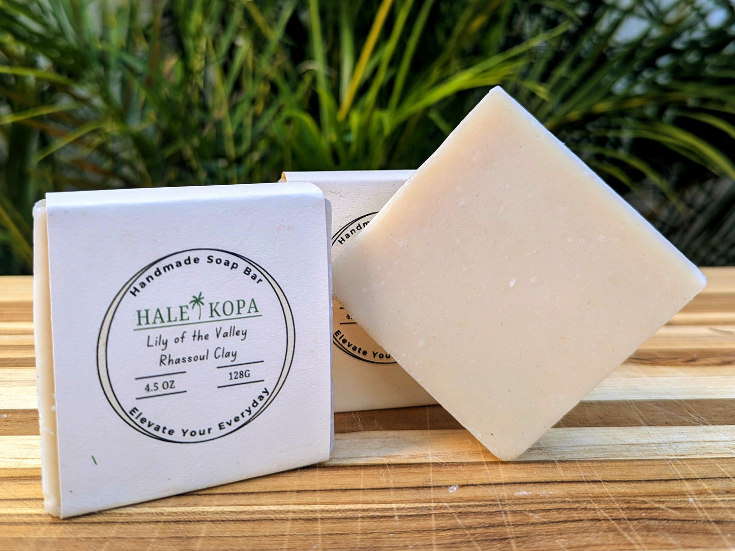 Two bars of soap with packaging that reads 'Lily of The Valley Rhassoul Clay', placed on a wooden surface with foliage in the background.