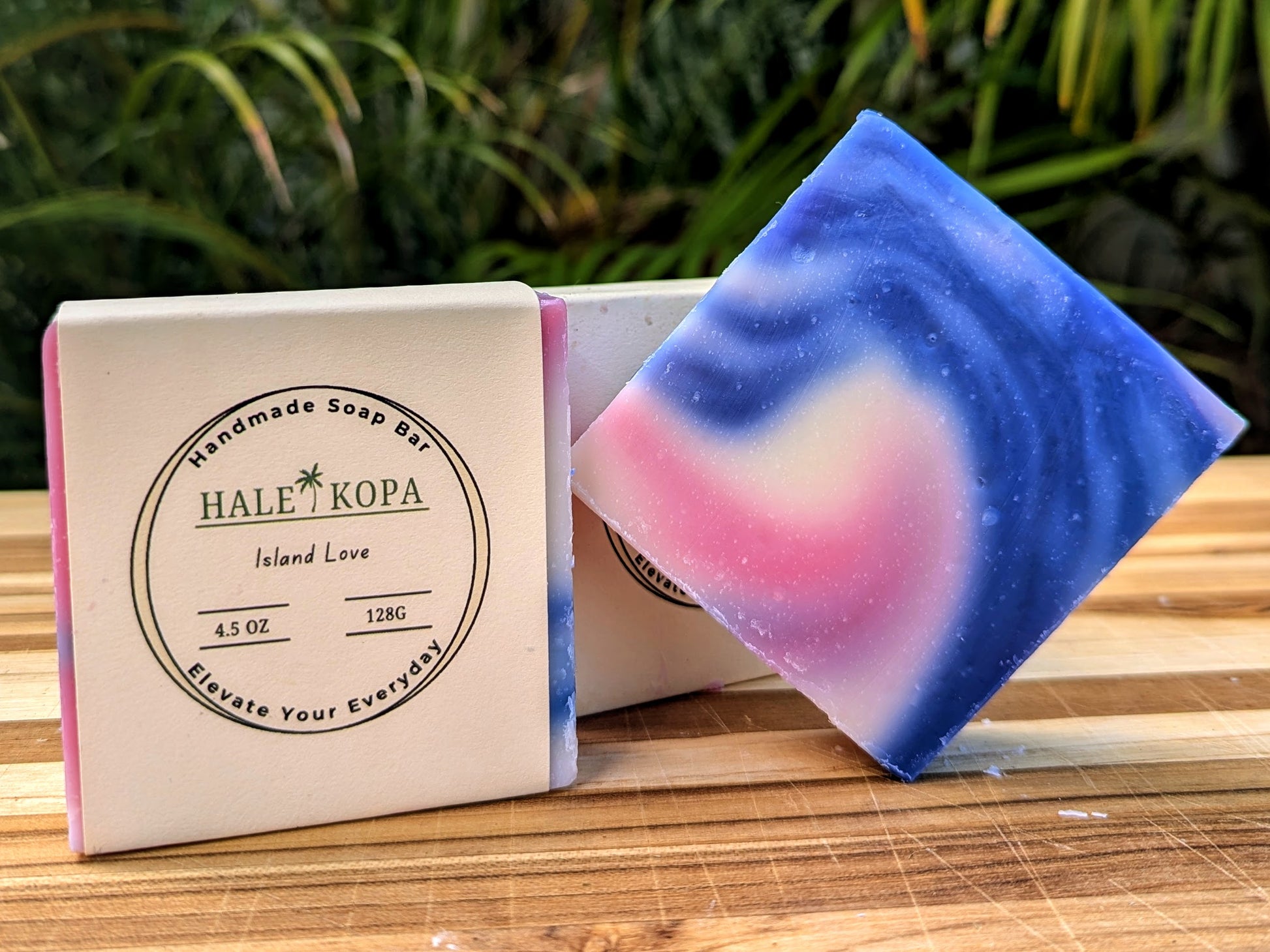 A bar of soap with a colorful design, featuring pink and blue hues, resting next to its packaging which reads 'Hale Kopa Island Love'.
